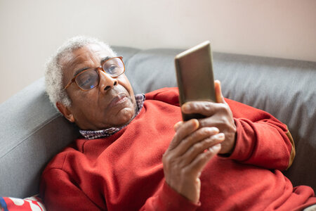 Elderly man with eyeglasses looking at his smartphone while laying on couch